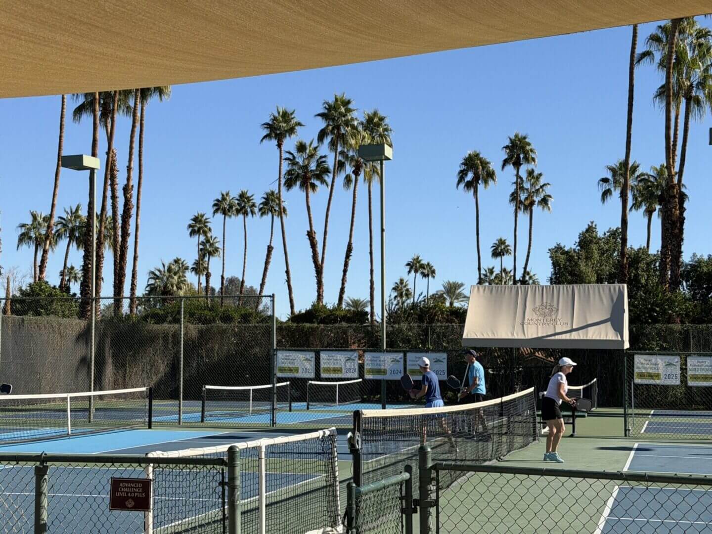 People playing tennis on an outdoor court with tall palm trees.